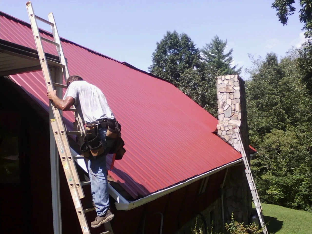 Roofer on a ladder beside a red metal roof and stone chimney — Soffit & Fascia Repair by Roofing Brothers in Massapequa Park