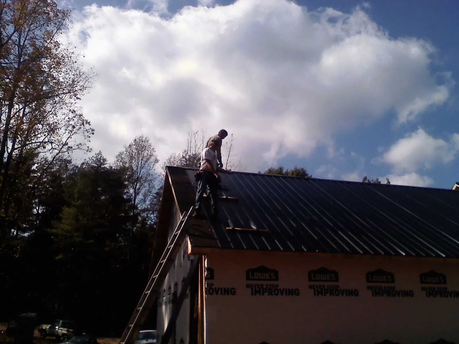 Roofer fastening dark metal panels high against a cloudy sky, from a Roof Inspection job in Massapequa Park